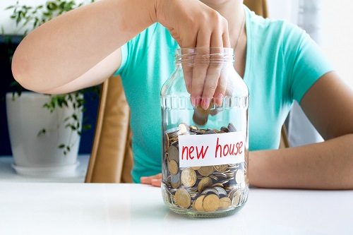 Closeup photo of young lady in blue T-shirt sitting at the white table on which is located ,full of coins , jar with the inscription new house